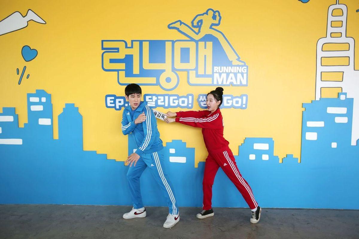 ชายหญิงสองคนในชุดกีฬา Running Man Experience Center ปูซาน ขณะกำลังสนุกกับกิจกรรม มีกำแพงป้ายซึ่งเป็นสัญลักษณ์ของ Running Man อยู่ด้านหลัง