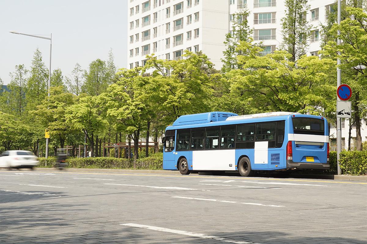 Bus kota berwarna biru di Korea, sedang berhenti di jalanan kota, dengan gedung apartemen putih dan pepohonan hijau di sekitarnya.