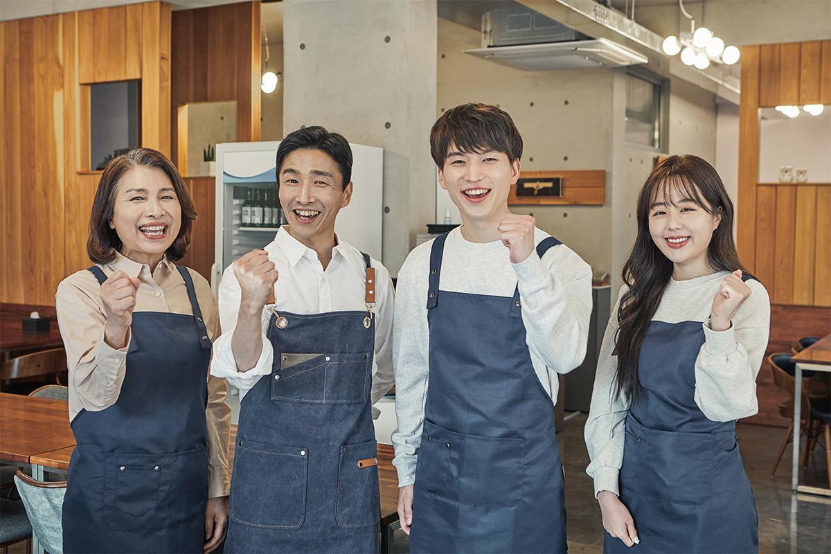 Nhóm sinh viên quốc tế mặc tạp dề xanh trong một quán cà phê ở Hàn Quốc, tay giơ lên thể hiện sự quyết tâm với bầu không khí làm việc hiện đại và thân thiện.