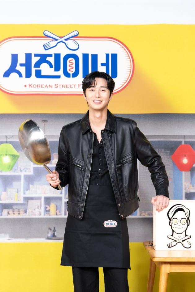 黒い革ジャンを着た人物が、大きなスプーンを持ちながら、「ソジンの家」のロゴ前でポーズを取っている。