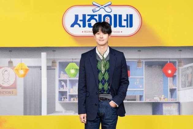 茶色いカーディガンを着た人物が、切られた牛肉のステーキ模様のファンを持ちながら、「ソジンの家」ロゴ前でポーズしている。