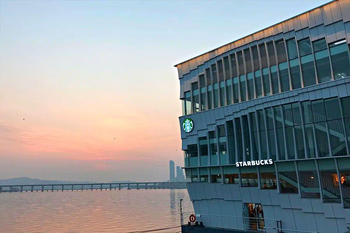 ภายใน Starbucks Seoul Wave Art Center พร้อมกับแสงไฟตกแต่งและเก้าอี้ที่ดูสวยงามลงตัว มองเห็นวิวแม่น้ำฮันและท้องฟ้าทางหน้าต่างใหญ่