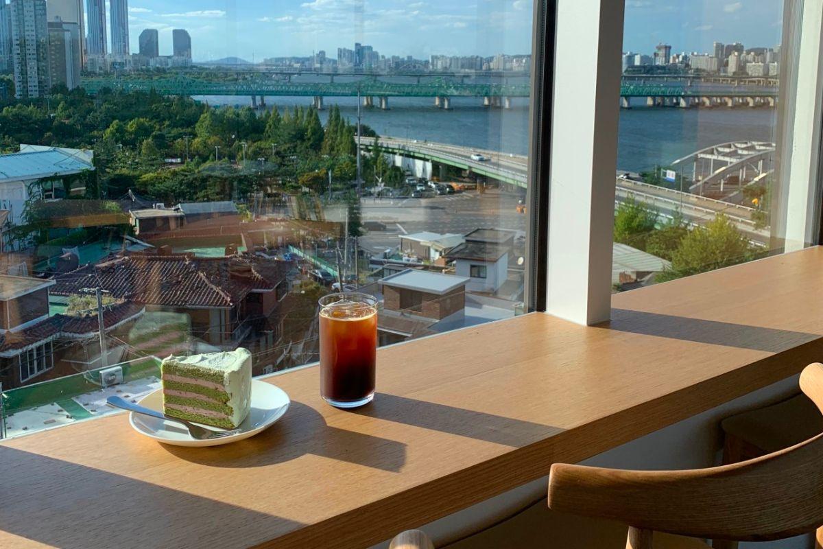 บรรยากาศภายในคาเฟ่ THE Hangang ที่มีหน้าต่างขนาดใหญ่ เปิดรับวิวของแม่น้ำฮันและสะพาน ซึ่งให้ความรู้สึกสงบและอบอุ่นในช่วงยามบ่าย