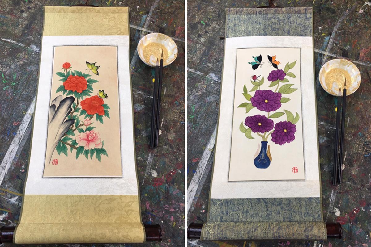 Dua lukisan gulung minhwa Korea tradisional dilengkapi dengan kuas di atas meja seni.