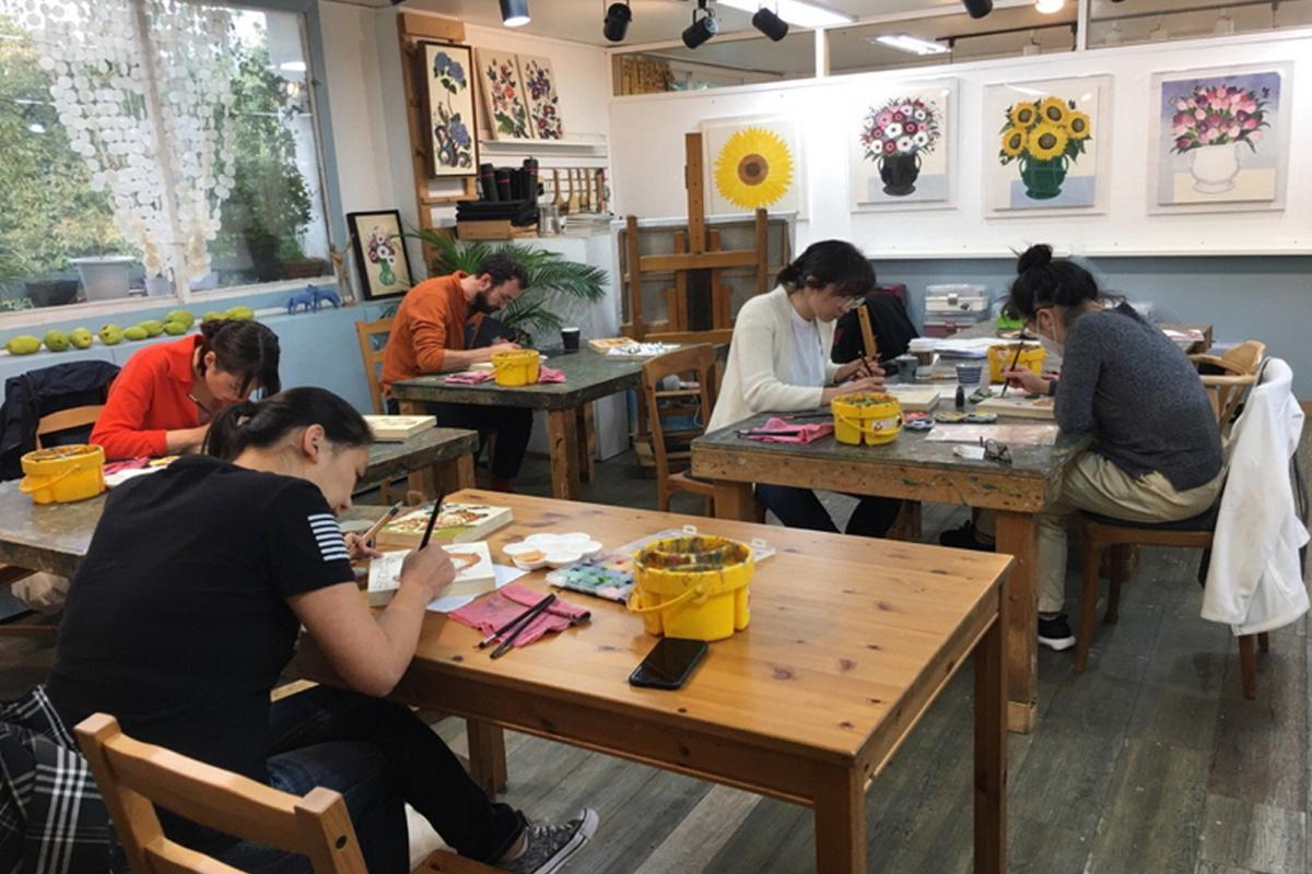 Para peserta sedang asyik menggambar seni minhwa di ruangan yang nyaman dan berdekorasi dengan lukisan bunga di dinding.