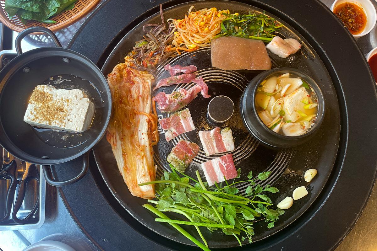 铁锅盖上摆放着泡菜、水芹菜等，与五花肉一同烤制，充分融合食材的风味