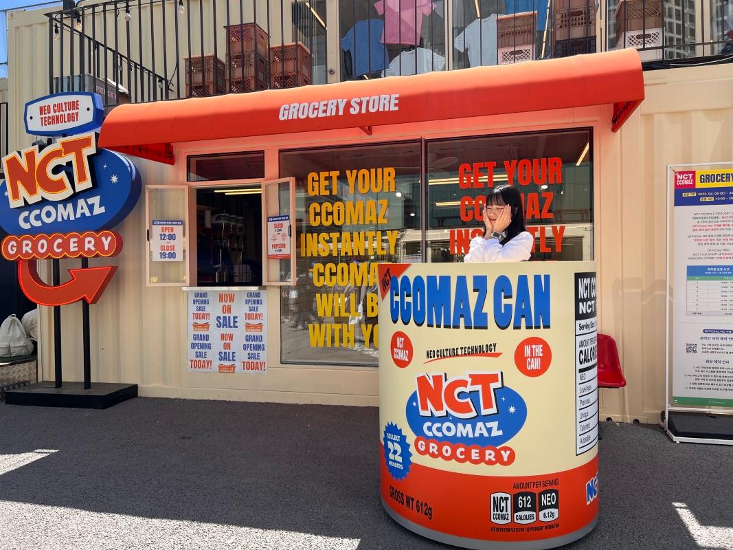 NCT CCOMAZ GROCERY STOREの外でNCTのCCOMAZキャラクターオブジェと撮影する訪問者。おしゃれで楽しいフォトスポット。
