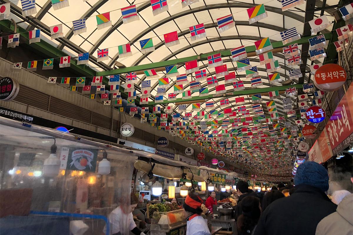 Jalan pasar yang ramai di Pasar Gwangjang, dihiasi dengan bendera internasional yang menggantung di atap.