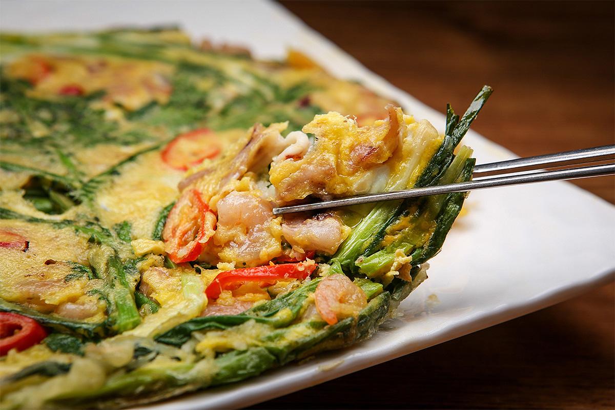 Pajeon coreano, un tipo de panqueque con cebollino y mariscos, mostrado en un primer plano con una porción levantada por un par de palillos.