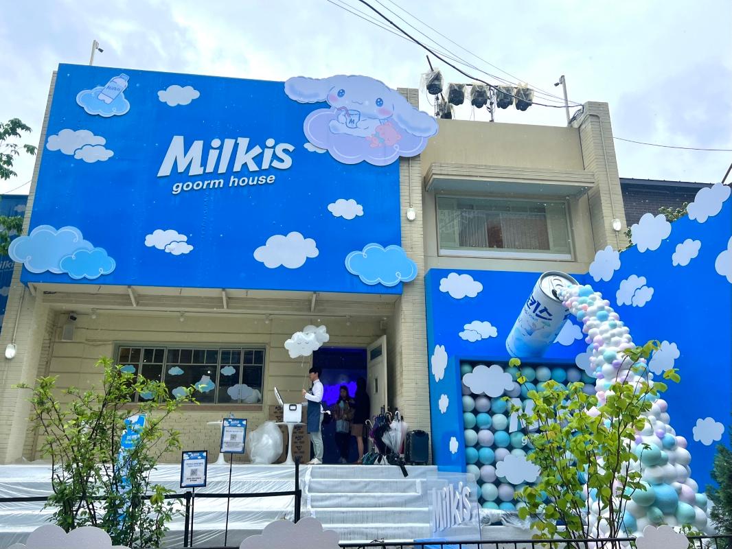 『Milkisゴームハウス』のカフェ外観です。青空と雲のデザインがプリントされており、シナモロールの可愛いイラストが目を引く看板が飾られています。