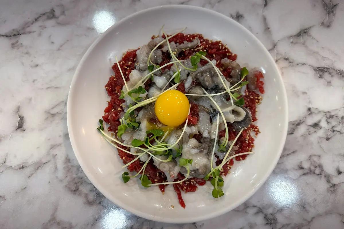 広蔵市場 昌信ユッケ 4号店の名物、韓国式ユッケタンタンイが白いお皿に盛られて、卵黄、野菜と一緒に飾られている。韓国旅行の際のおすすめ料理で、ユッケと生タコの組み合わせが楽しめる一品です。SEO対策のための最適なALTテキスト。