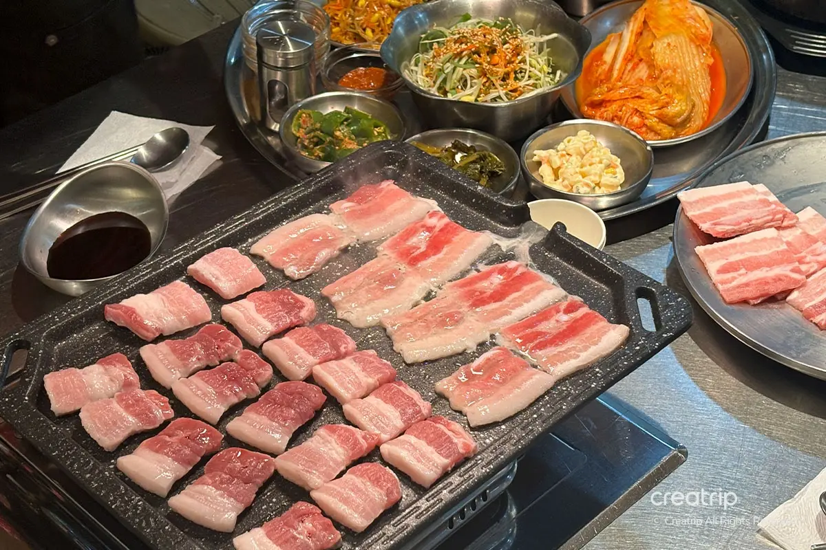 坊豚食堂内正在烧烤的生三层肉，展示传统的韩国烧烤风格，配有多种美味的韩式小菜。