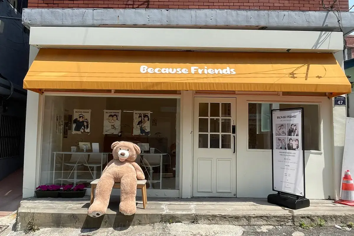 Exterior of Because Friends photo studio in Busan, featuring a large teddy bear on a chair under a yellow awning.