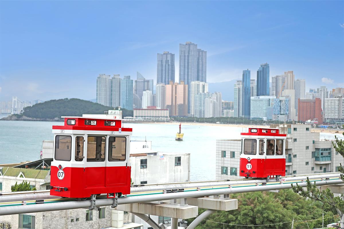 Красный и белый пляжный поезд Blue Line, движущийся вдоль побережья Пусана с видом на море и город.