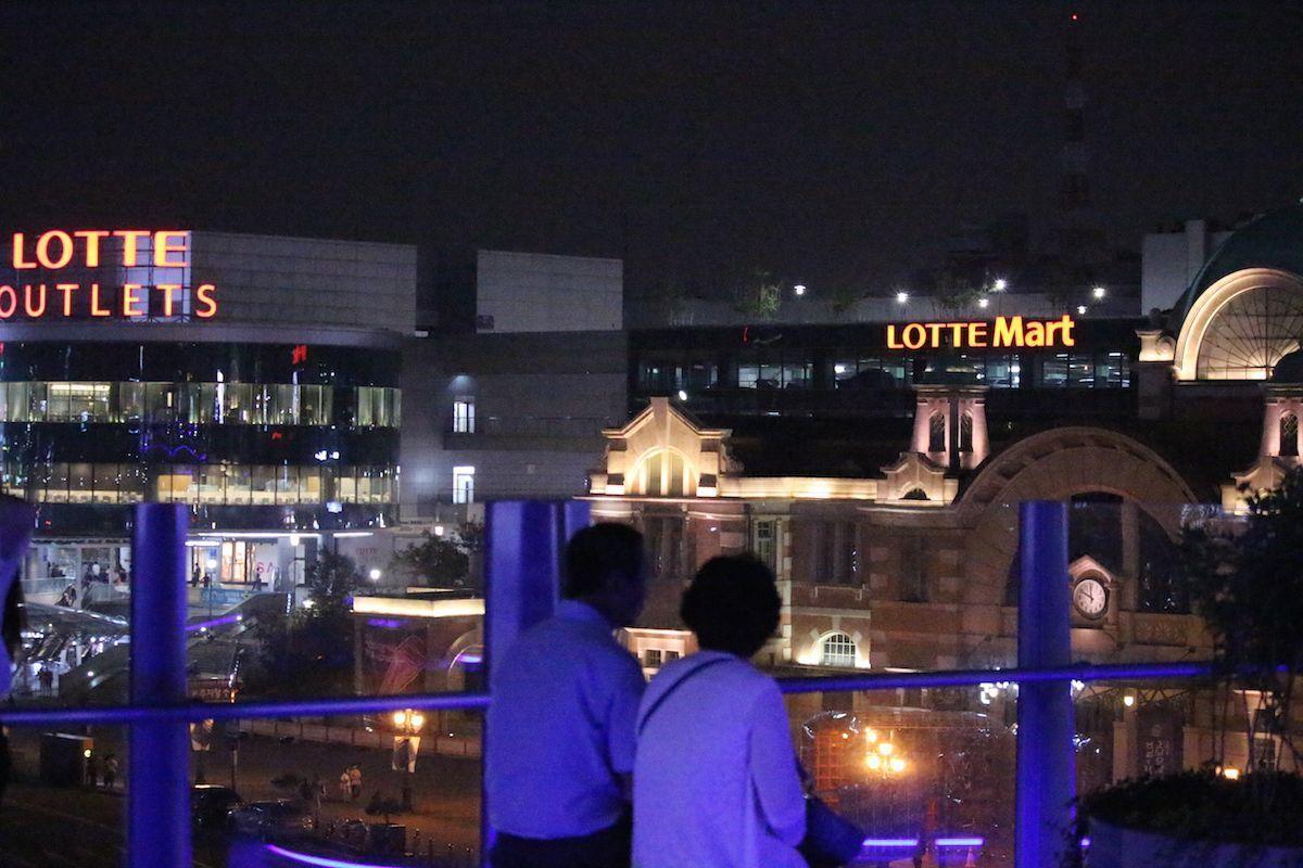 Couple overlooking the nighttime view of Lotte Mart and old Seoul Station, captured from Seoullo 7017.