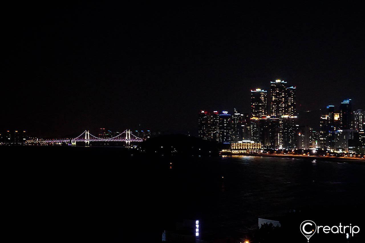 Night view of Gwangan Bridge and Haeundae beach in Busan, illuminated by city lights and reflections.