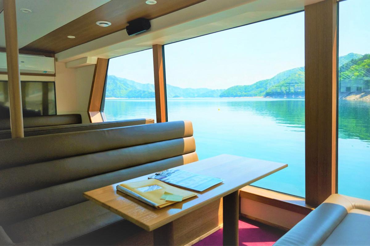 Энх тайван Нури аяллын дотор талын зураг. Их байрны дотор тал аятайхан суух буйдан ба ширээнээр хүрээлэгдэнэ. Түргэн амралтанд хүрэх боломжтой горхи оройн харгейсээр харуулж буй хөлгөн дээрээс харагдаж байгаа.