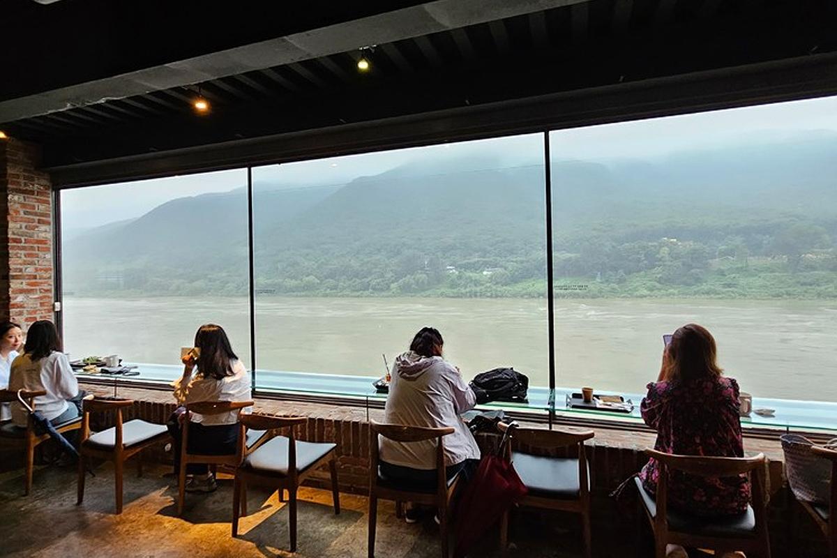 Cafe patrons enjoying a serene view of a river and mountains from inside a popular cafe in Korea.