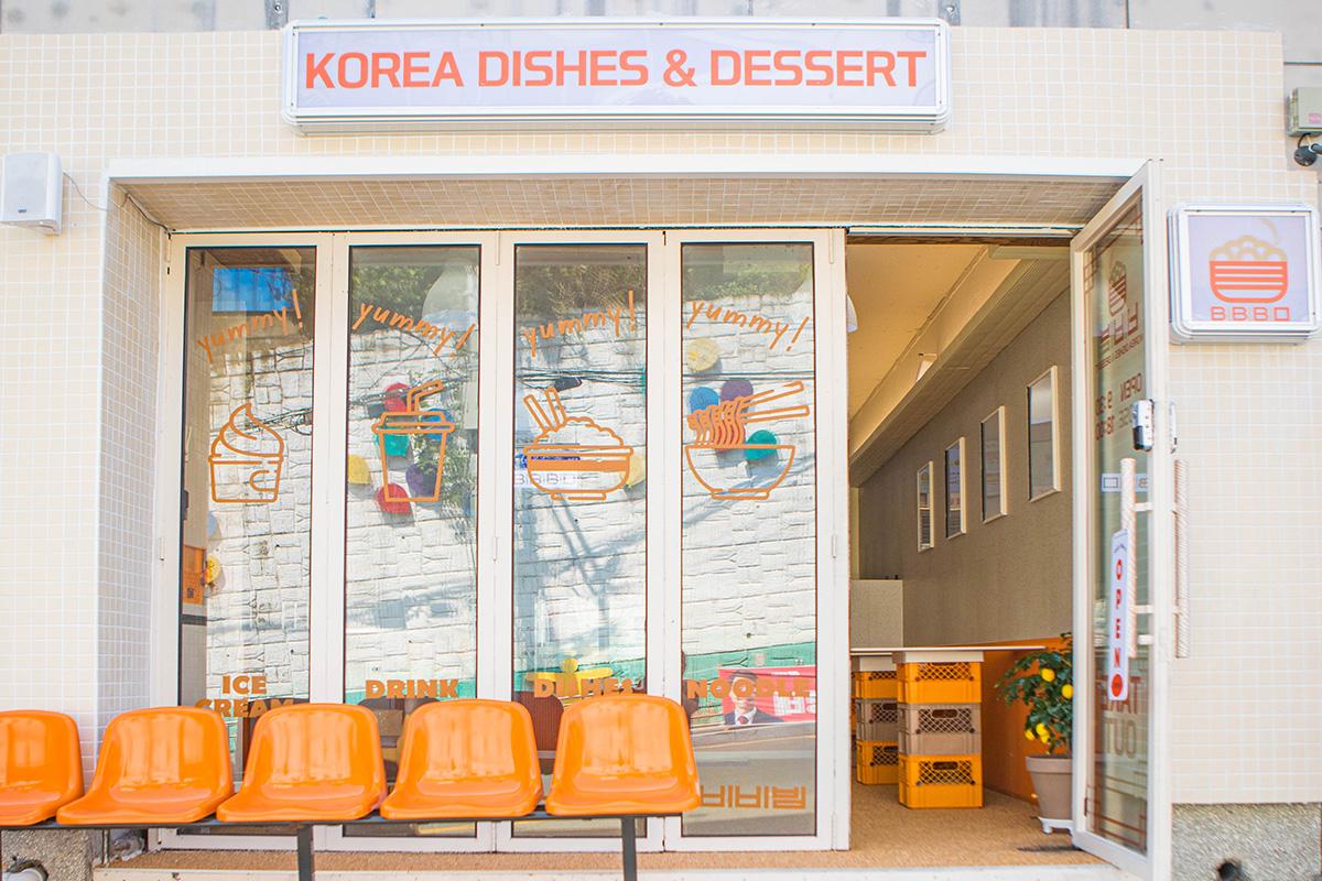 Exterior view of BIBIBIM cafe and restaurant located near Gamcheon Culture Village in Busan, showcasing its lively white and orange theme and a sign that reads 'Korea Dishes & Dessert'.