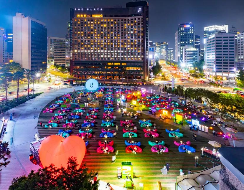 首爾市中心夜間廣場活動場景，成排彩色充氣懶人沙發與燈光裝置，遊客在戶外享受初夏晚間市集與文化活動，背景可見飯店與城市燈海。