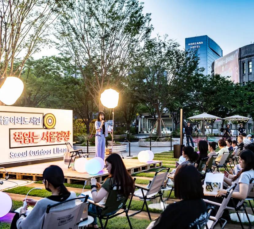 首爾戶外圖書館活動傍晚場景：舞台上的講者在柔和打光下演講，前方觀眾坐在折疊椅上聆聽，周圍樹木與城市建築形成初夏悠閒氛圍，適合介紹6月首爾戶外閱讀與晚間活動建議。