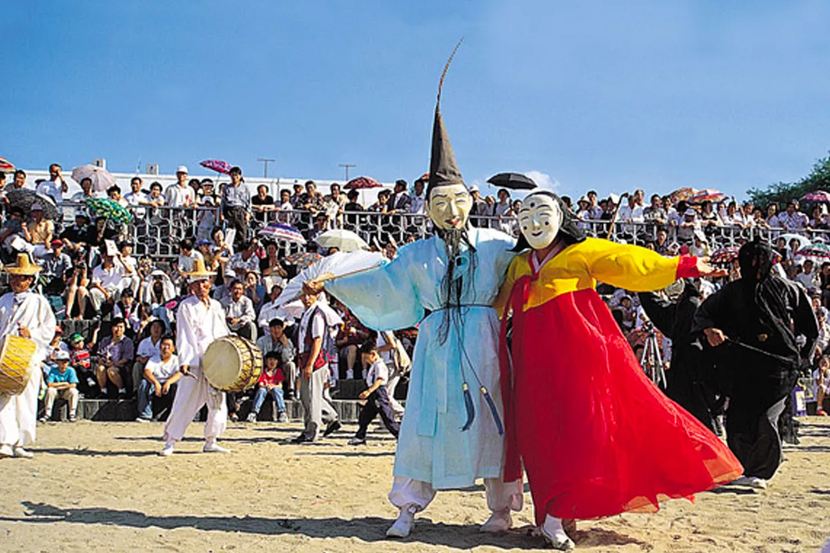 江陵端午祭街頭場景：穿著傳統服飾的表演者與鼓手在沙地上遊行，後方看台坐滿觀眾撐陽傘，呈現節慶熱鬧氛圍與民俗活動特色。
