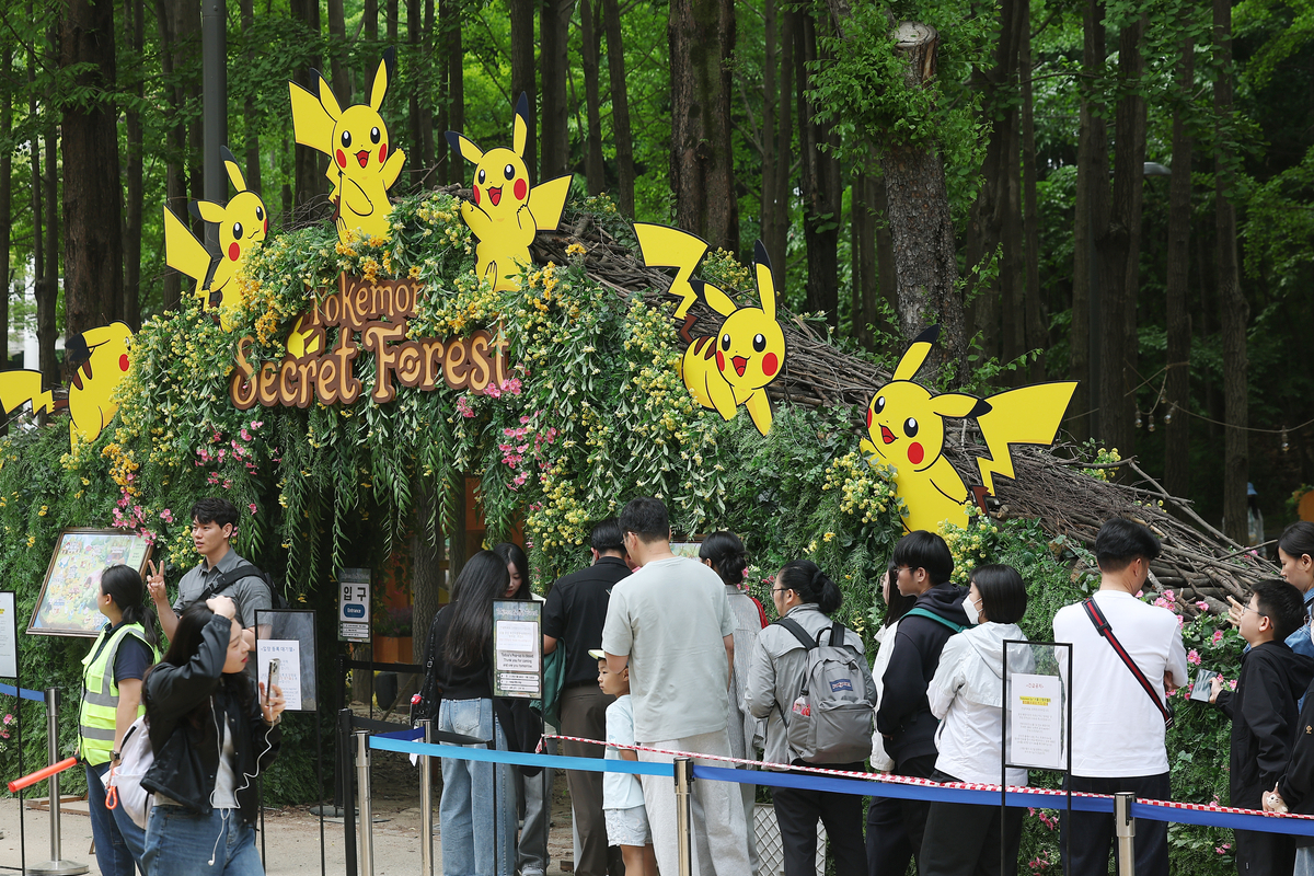 首爾林間展區排隊入場情景，入口以綠植與皮卡丘裝飾標示「Pokemon Secret Forest」，遊客穿著初夏服裝，手持雨具與背包，適合介紹6月觀展與穿搭建議。