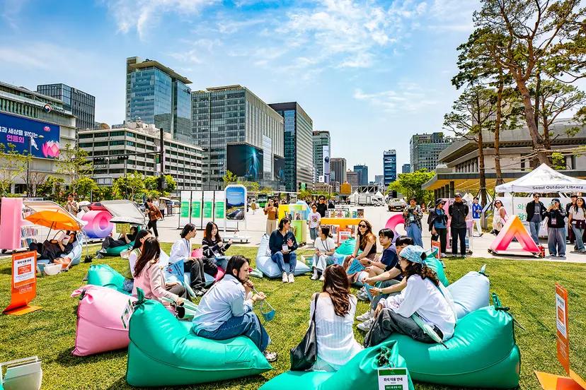 首爾市中心戶外圖書館活動現場，晴朗初夏午後多人坐在彩色充氣懶人沙發上閱讀與聊天，背景可見高樓與臨時展攤，呈現6月戶外休閒與文化市集氛圍。