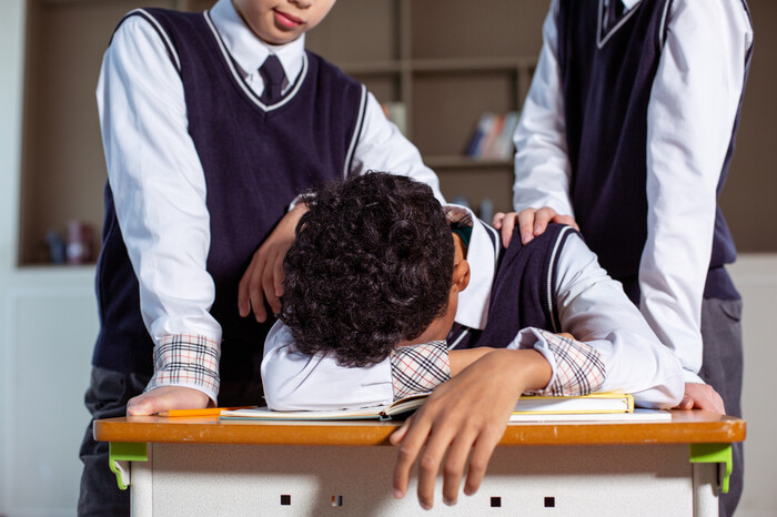 韓國學校教室內部：一名學生趴在桌上，兩名同學站在他身邊，一人用手接觸他的肩膀，顯示出可能的欺負情況。