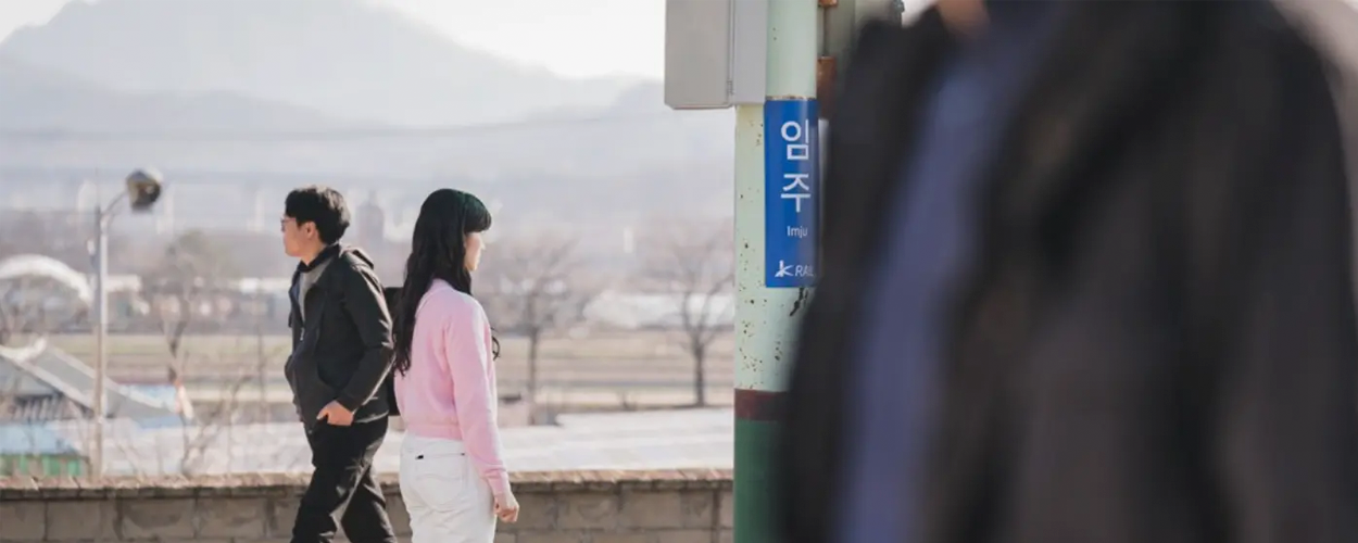 Scene from Lovely Runner filmed at Imsil Station, featuring actress in pink sweater beside an Imju signpost, with mountains visible in the background.