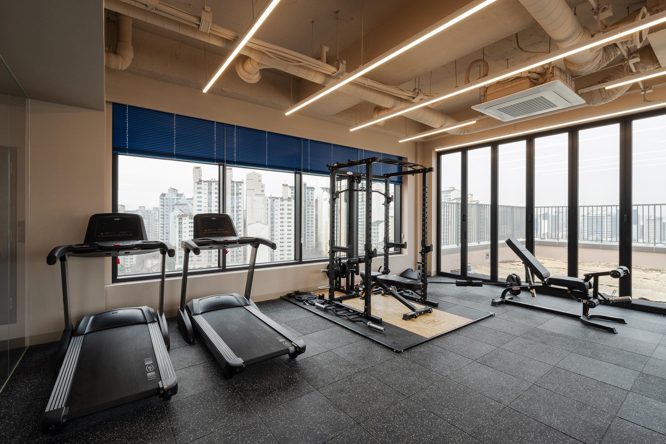 ห้องออกกำลังกายใน Co-living House Mangrove Sinchon พร้อมกับอุปกรณ์ออกกำลังกายพื้นฐานและหน้าต่างขนาดใหญ่ที่เปิดให้เห็นวิวเมือง