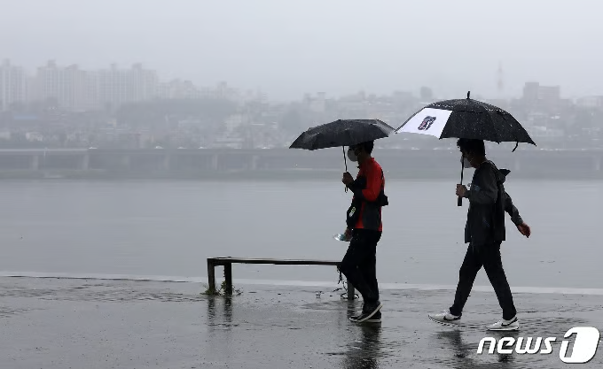 兩人撐著雨傘沿河岸行走，灰濛濛的城市天際線在雨中隱約可見，呈現韓國梅雨季常見的陰雨與潮濕氛圍。
