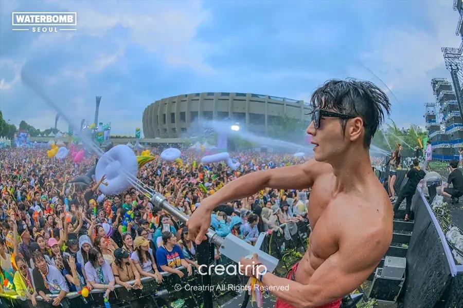 首爾夏日Waterbomb戶外水上派對場景：肌肉男用水槍向歡呼的人群噴水，午後多雲但氣氛熱烈，適合提醒旅客備防曬與備雨用品。