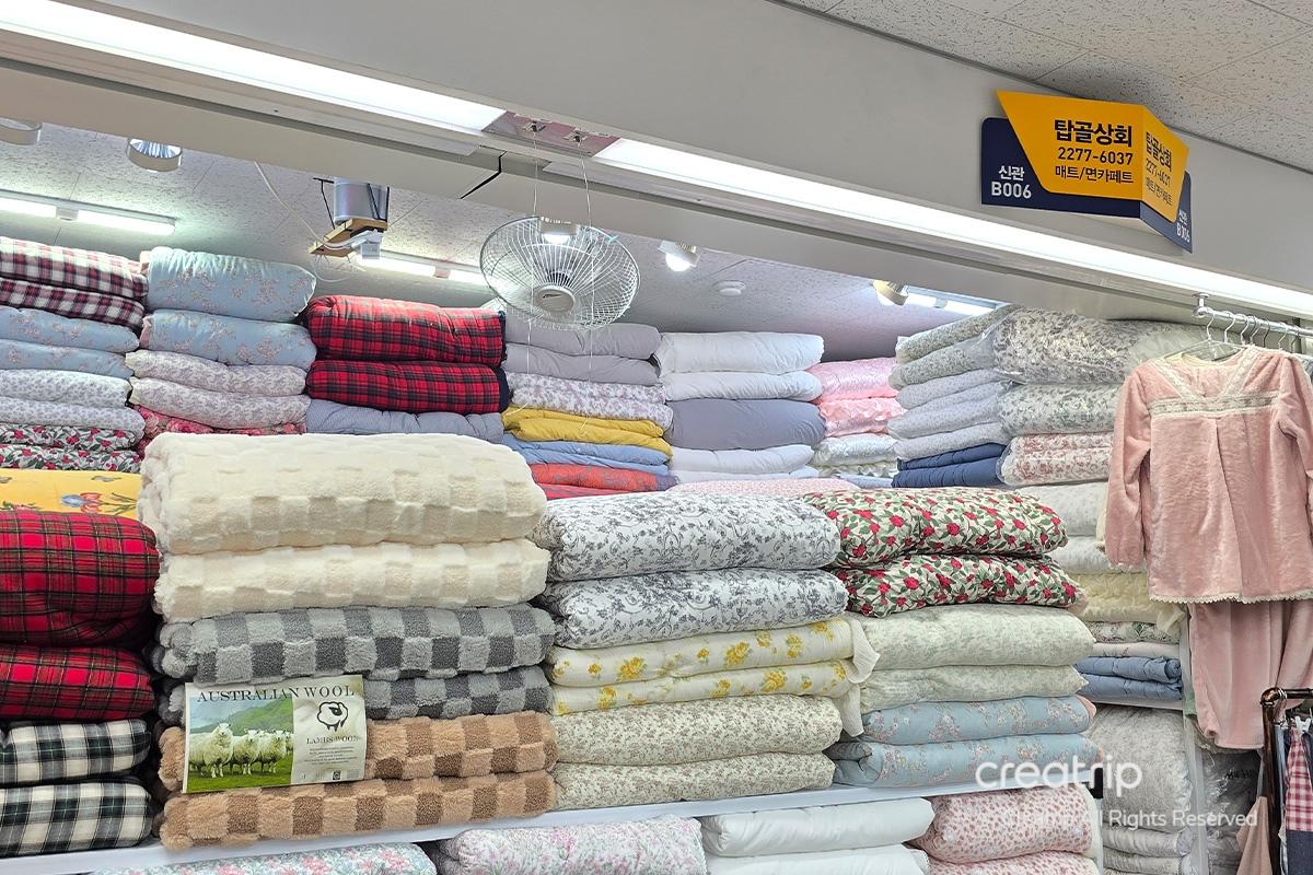 塔洞商會店內各式印花被子和牀單整齊堆放，色彩繽紛，懸掛的牌子上標示電話號碼和店鋪信息。