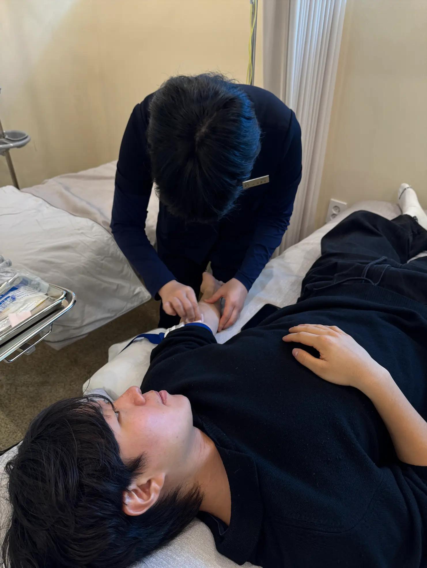 クリニックの半個室で横になり点滴の準備を受ける男性患者。スタッフが腕に包帯や固定具を巻いて処置しており、隣に点滴セットや医療トレイが置かれている。リラックスして横たわる様子が写っている。