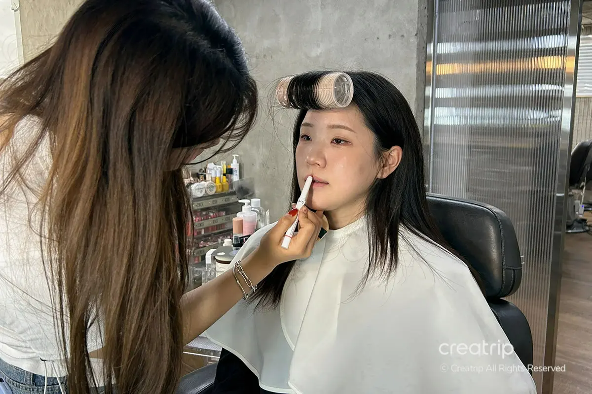 Makeup artist applying lipstick during an idol-style makeover in a salon: stylist leans in to apply color while client wears a white cape and hair roller on forehead.