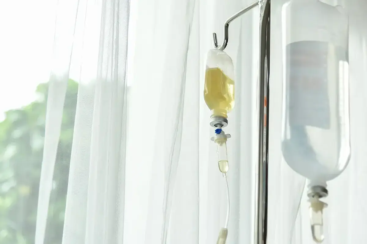 IV vitamin therapy setup: two intravenous fluid bags hanging from a metal stand beside a window with sheer curtains, soft natural light.