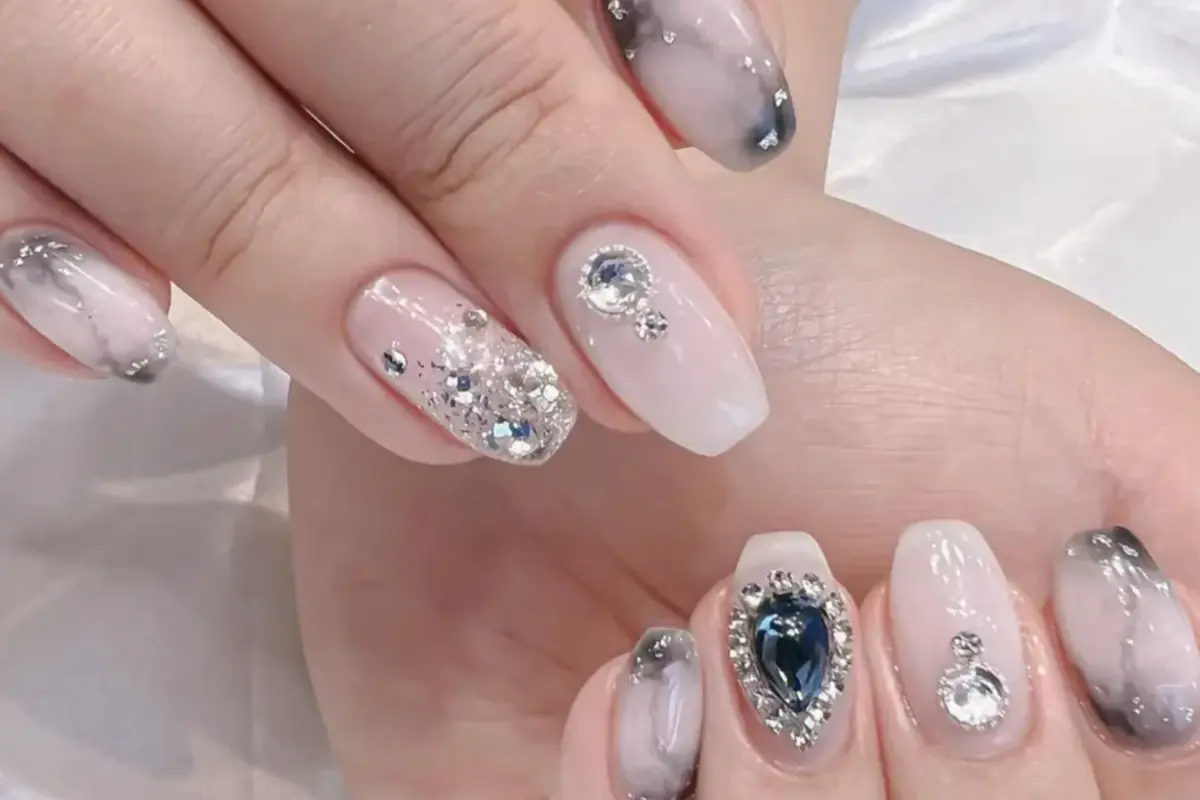 Close-up of premium nail art featuring pale pink and grey marble designs with glitter and rhinestone accents, including a teardrop-shaped blue gem on one nail.