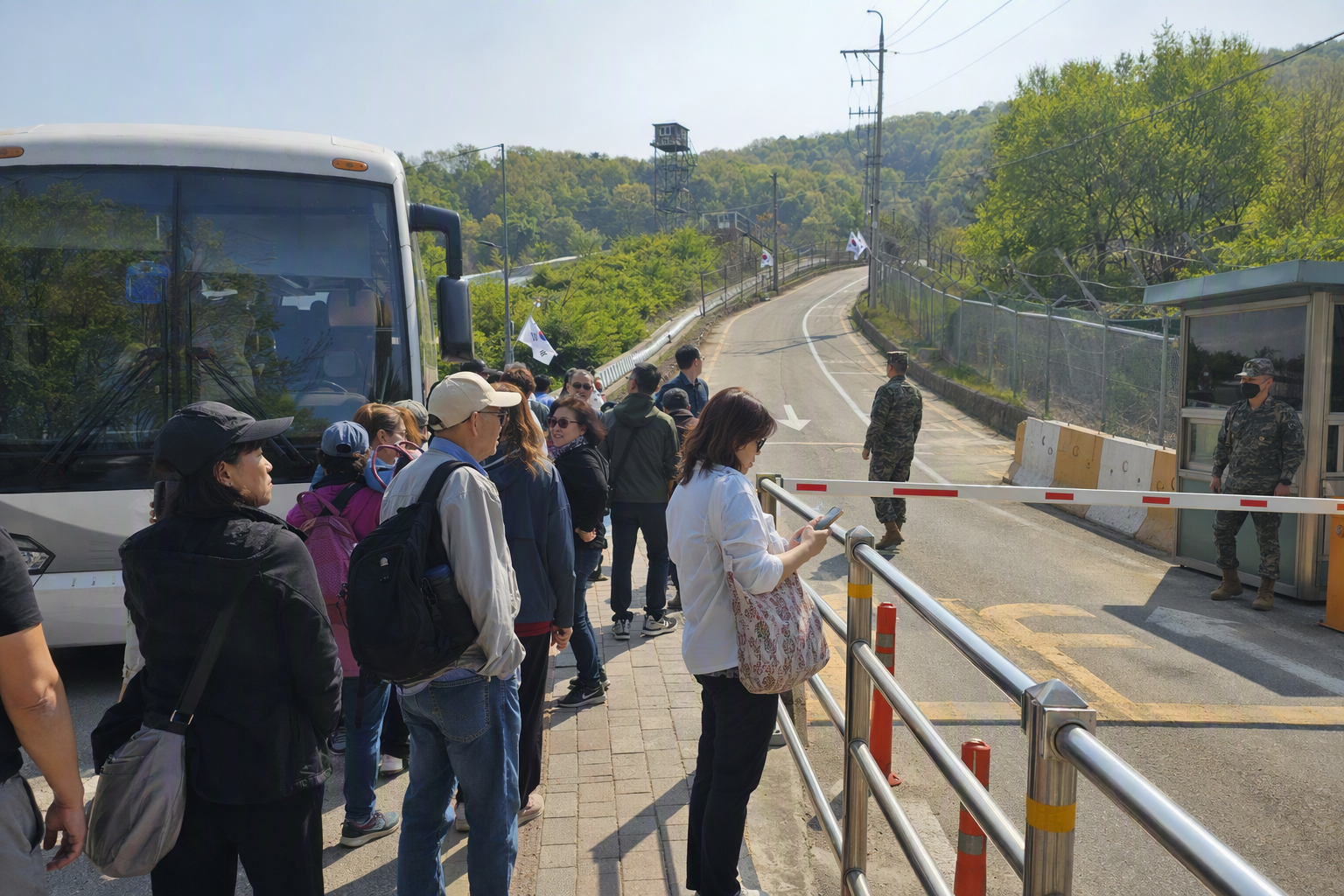 Photorealistic DMZ tour bus near a controlled checkpoint road, travelers waiting, security barriers, sunny day, realistic travel scene, no Korean text