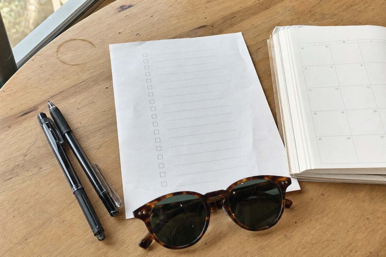 Flat lay of a simple travel checklist with a pen, sunglasses, and a blank calendar page, bright and clean, photorealistic, no text