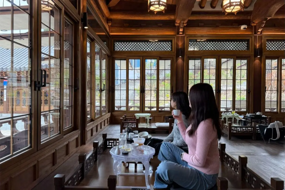 Two women sipping tea at a low table inside a sunlit hanok room, looking out through large wooden-framed windows to a courtyard.