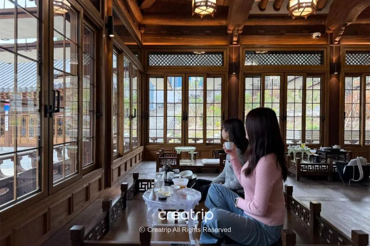 首爾忠武路韓屋茶室內景，兩位旅客靠窗坐在傳統低桌前慢品茶點，窗外庭園與木構架梁柱營造靜謐韓屋氛圍，適合體驗宮廷茶果。
