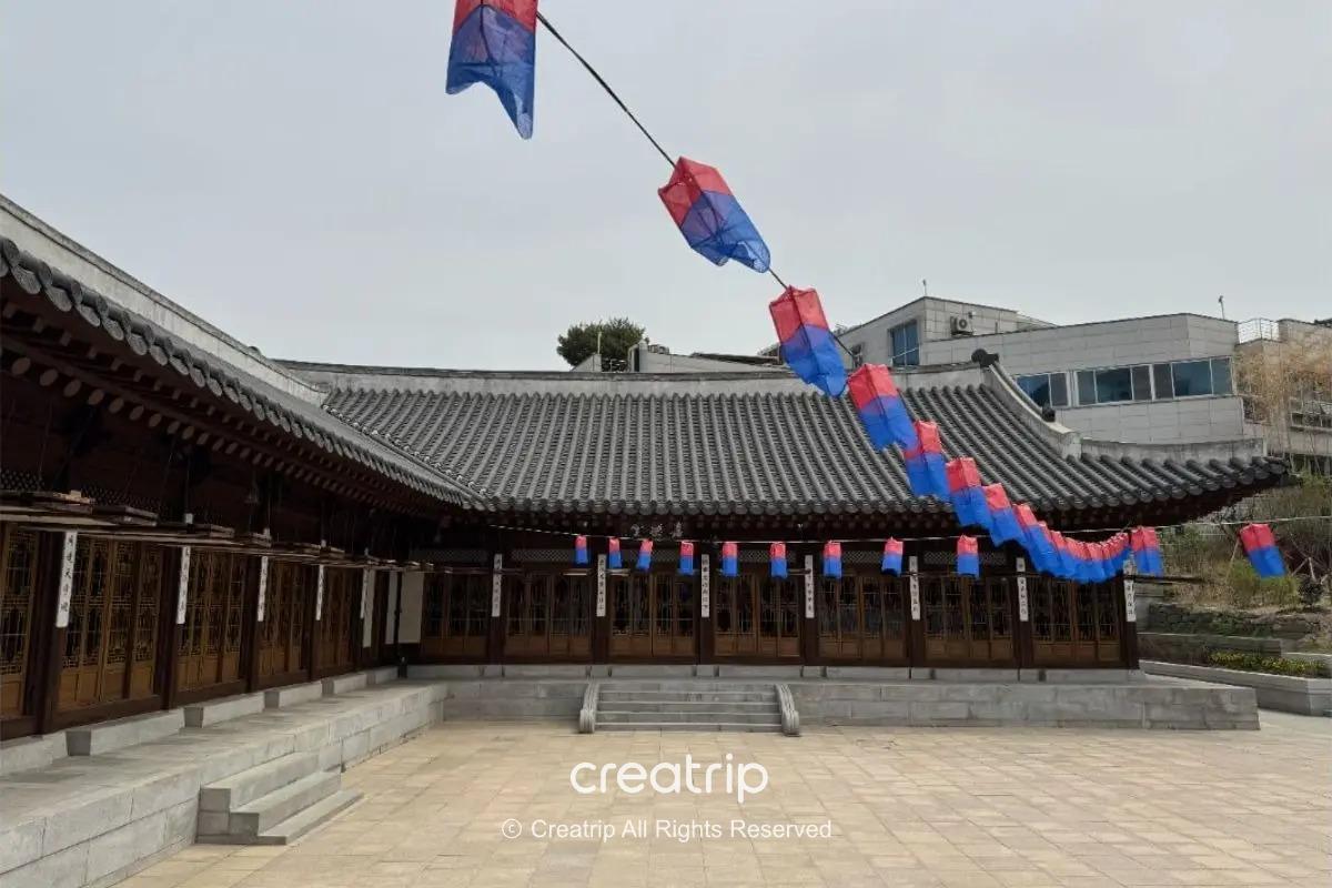 首爾市中心的韓屋院落外觀，瓦屋簷與木製窗扇環繞，庭院石階與廣場鋪面，紅藍燈籠掛成一排，呈現古好齋沉穩雅致的傳統氛圍。