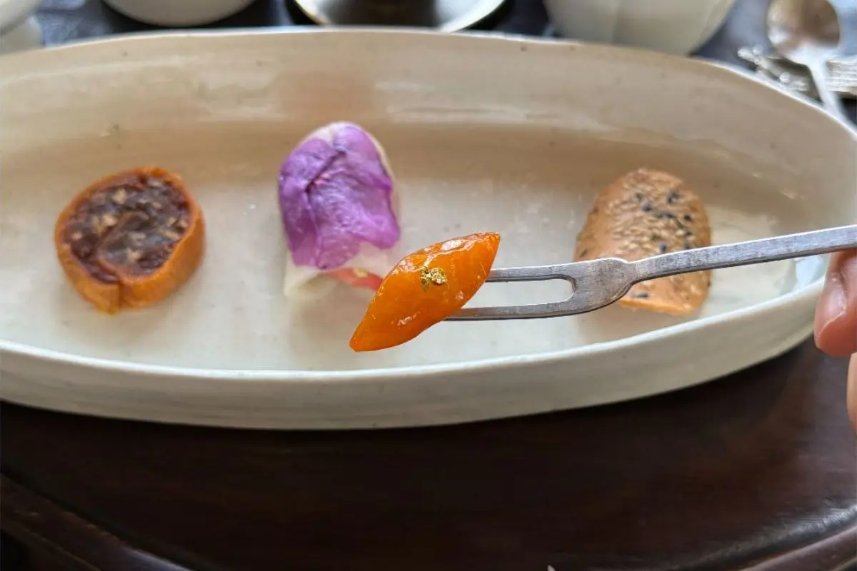 一人份韓式茶點盤近拍：白色長盤上排列烤年糕、花瓣裝飾點心與芝麻煎餅，前景小叉子夾起帶金箔的蜜釀金桔，呈現古好齋季節傳統甜點與精緻器皿的靜謐氛圍。