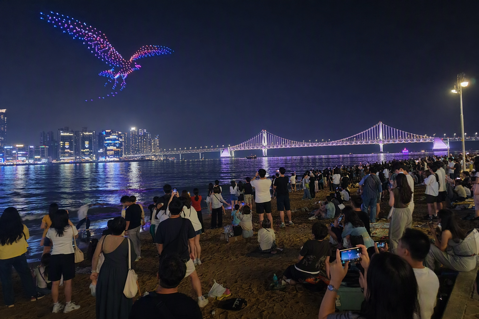 Vibrant photorealistic night shot of a drone light show above a beach, illuminated bridge in the background, crowd silhouettes, glittering reflections