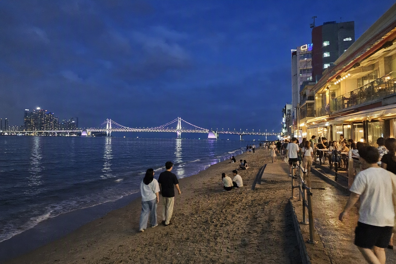 Bright vibrant photorealistic evening scene at Gwangalli Beach with illuminated bridge, people walking along the sand, lively cafes, deep blue sky, no text