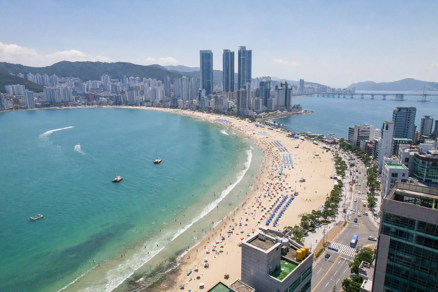 Bright vibrant photorealistic wide view of Busan coastline with blue sea, sandy beach, high-rise skyline, mountains in the background, sunny clear day, no text
