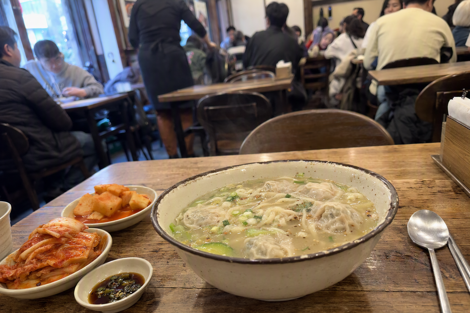 明亮鮮豔、寫實風格的一碗刀切麵湯，附餃子、湯頭、蔬菜切片與泡菜，簡單餐桌上，無可見文字