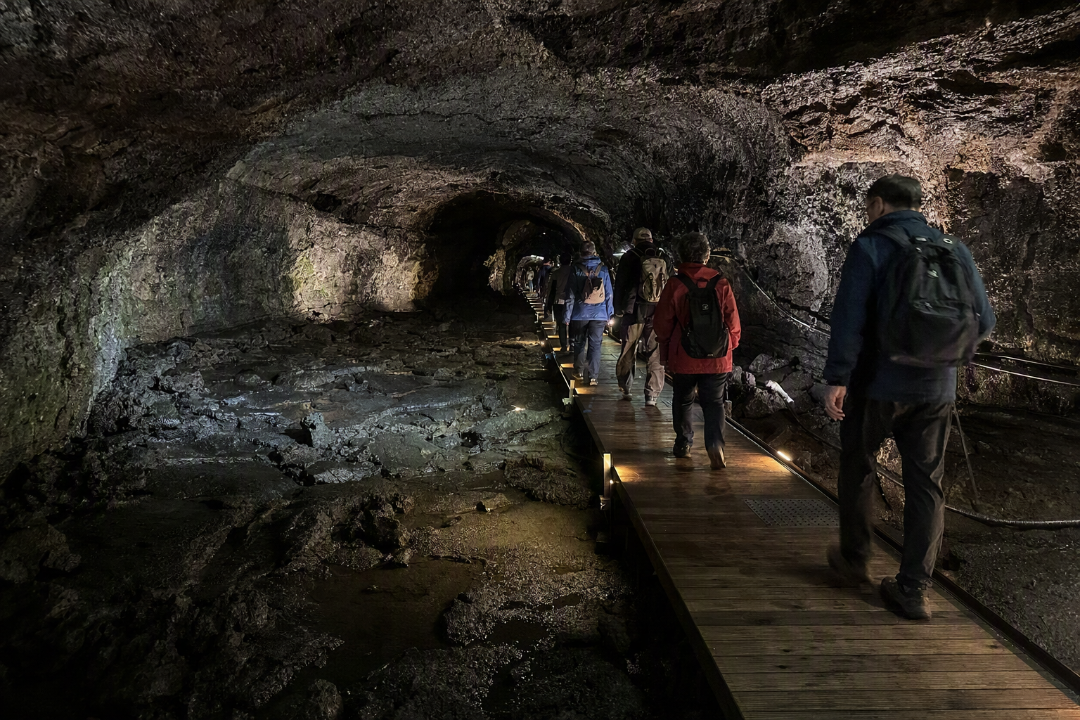 Bright photorealistic interior of a lava tube cave with dark volcanic walls, subtle pathway lighting, damp stone floor, and visitors in jackets, no text