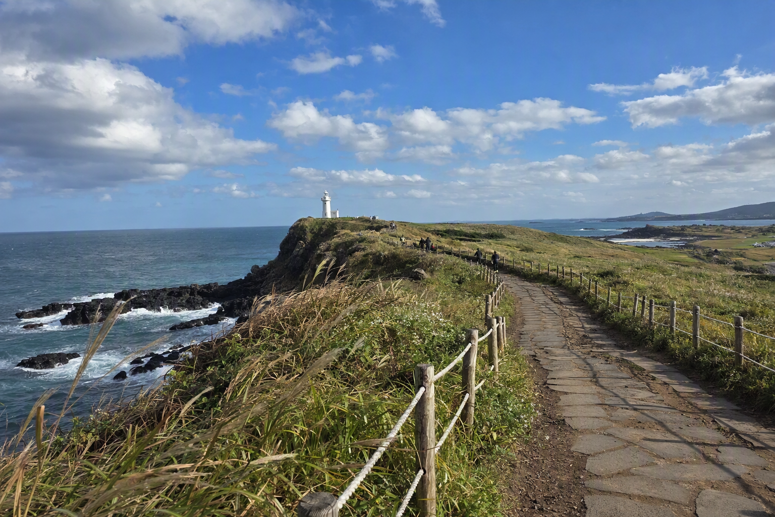 Bright photorealistic coastal trail at Seopjikoji with green grass, black rocks, blue ocean, white lighthouse, and dramatic clouds, no text